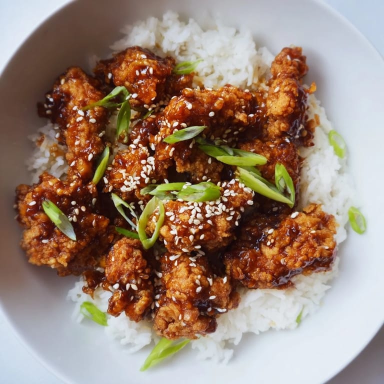 A delicious bowl of steaming, saucy General Tso's Chicken with fluffy white rice topped with sesame seeds.