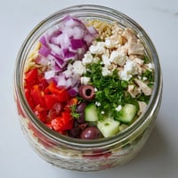 Clear glass mason jar layered with Greek Pasta Chicken Salad, showing colorful orzo, diced chicken, tomatoes, cucumbers, and crumbled feta.  